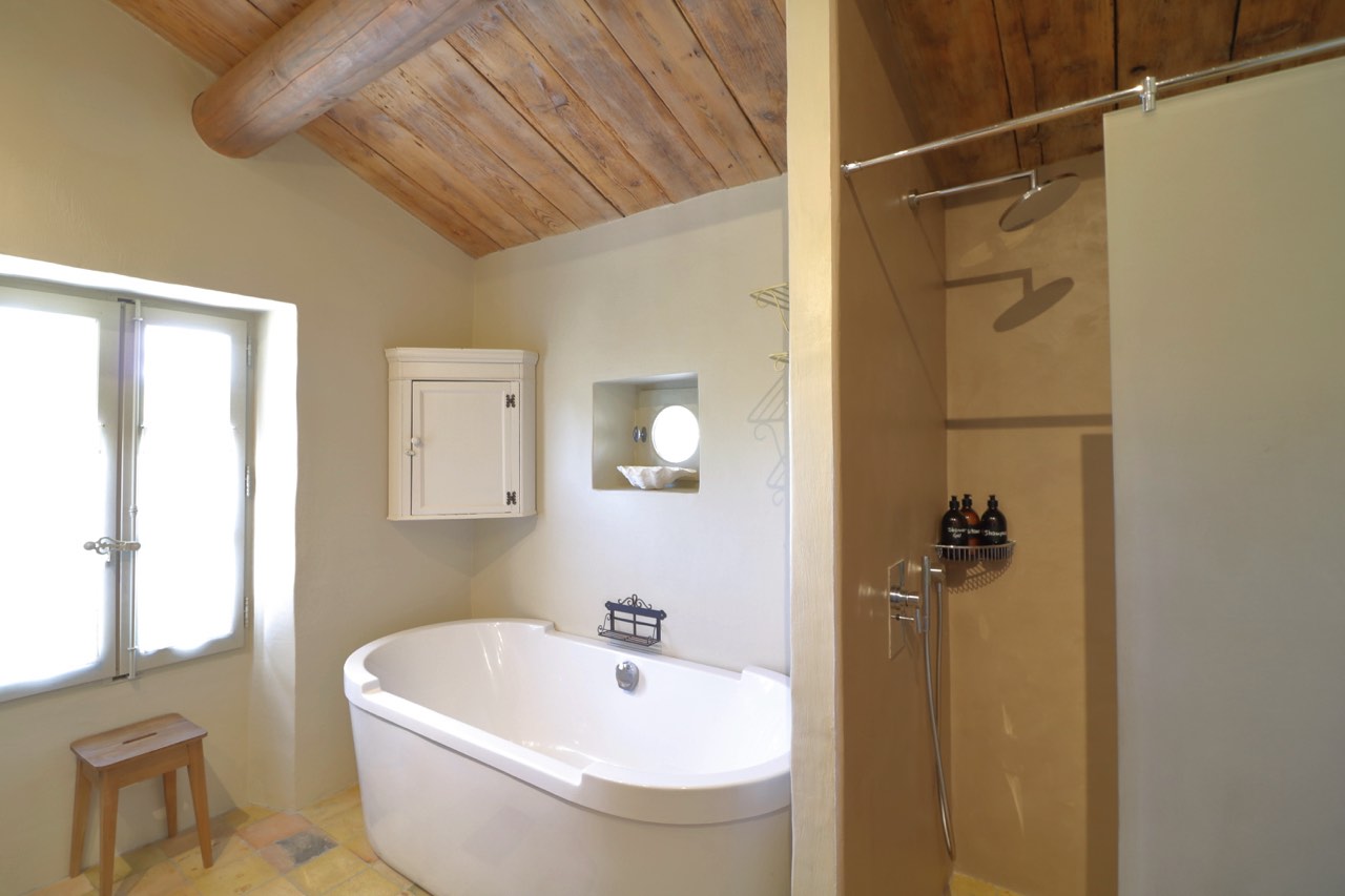 A bright and airy bathroom featuring a freestanding white bathtub, wooden exposed beams on the ceiling, a separate shower area, and a small, open window.